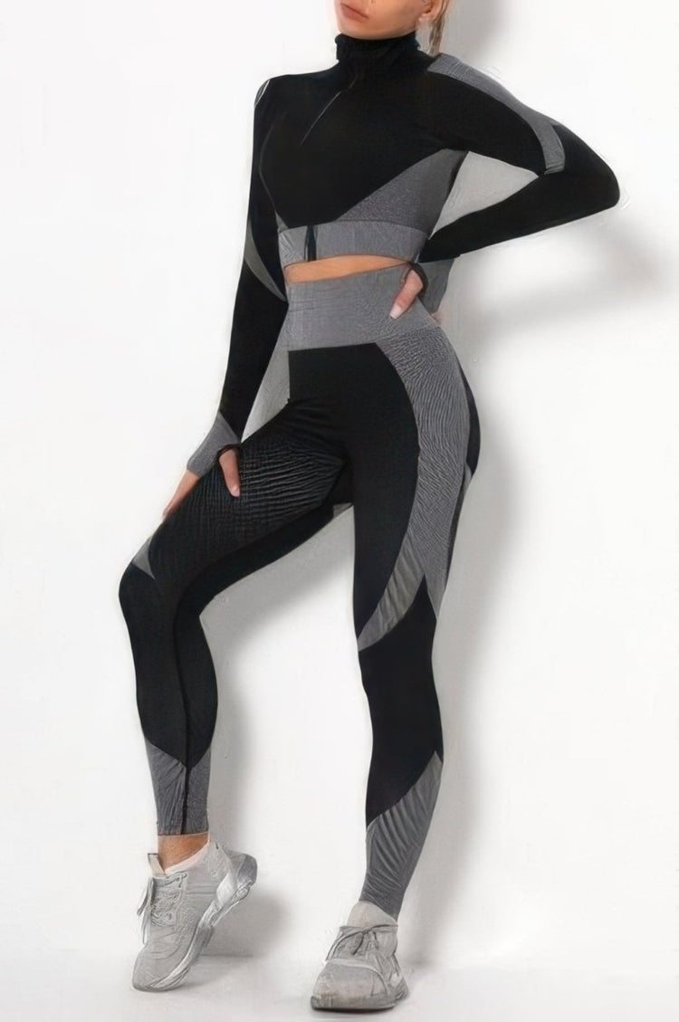 A woman modeling a matching two-piece athletic set consisting of a long-sleeve mock-neck crop top and high-waisted leggings, both in black and gray color-blocked panels, paired with gray sneakers.