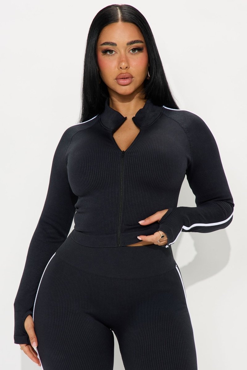 A woman modeling a black ribbed long-sleeve athletic top with a split neckline and white stripe accents on the shoulders and cuffs, paired with matching black leggings against a light gray studio background.