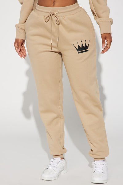 A person wearing beige drawstring sweatpants with a black crown graphic on the left thigh, paired with white sneakers against a light gray studio background.