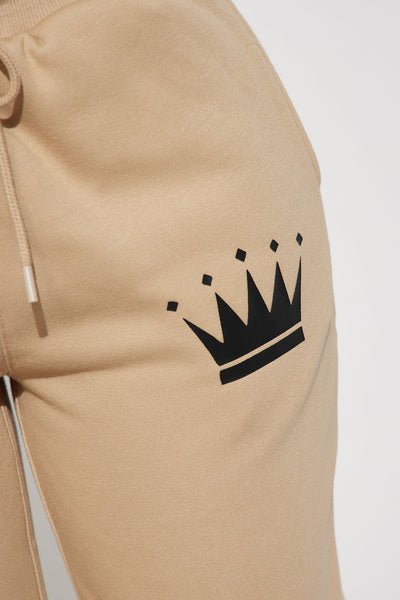 Close-up of beige drawstring sweatpants featuring a black crown graphic printed on the thigh.