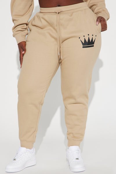 Beige sweatpants with a black crown logo worn by a person on a white background