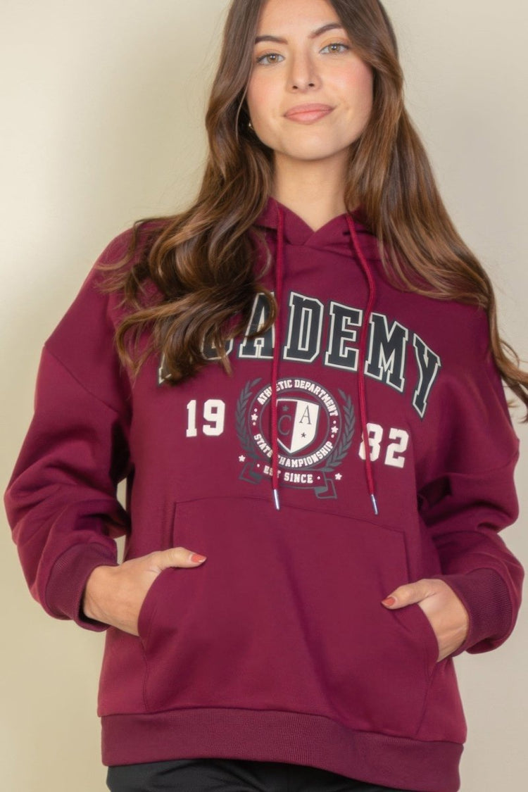 Woman wearing a maroon hoodie with 'Academy' text and design on a beige background