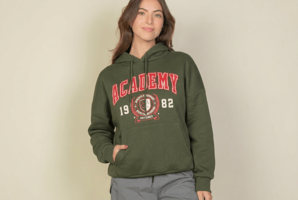 A woman wearing an olive green pullover hoodie with a red and white 'Academy 1982' graphic on the front, paired with gray pants, standing against a plain beige background.