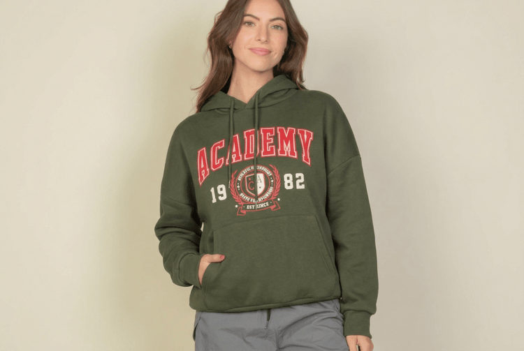A woman wearing an olive green pullover hoodie with a red and white 'Academy 1982' graphic on the front, paired with gray pants, standing against a plain beige background.
