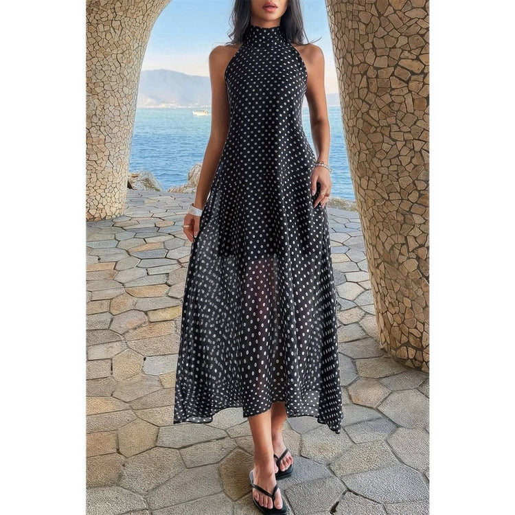 A woman wearing a sleeveless black midi dress with a white polka dot pattern, standing on a stone walkway between two stone pillars with a scenic ocean view in the background.