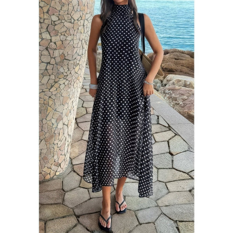 A woman wearing a sleeveless black midi dress with a white polka dot pattern, standing on a stone path with a stone pillar and ocean background.