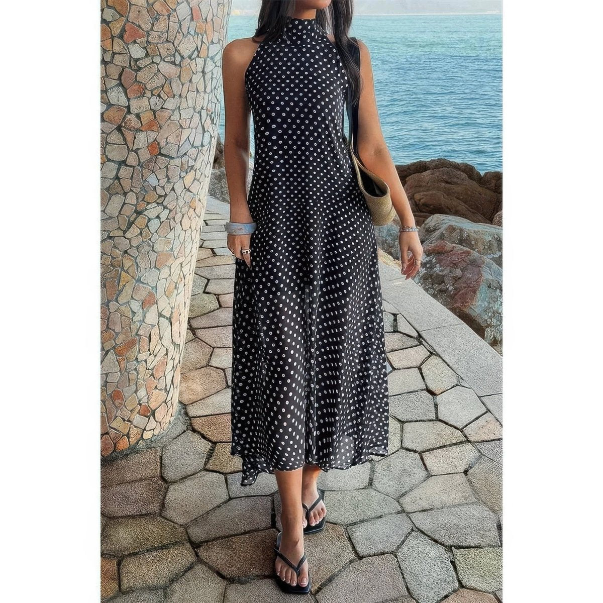 Woman in a black polka dot dress standing by a stone wall with ocean view