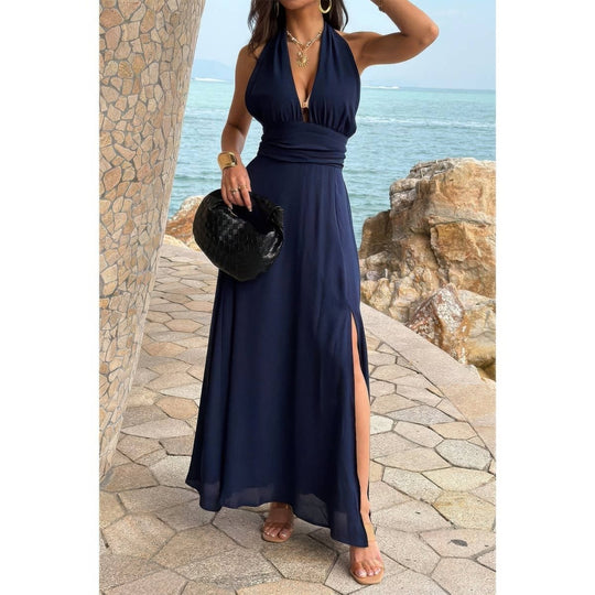 Woman in a navy blue halter neck dress standing by a stone wall with ocean view.