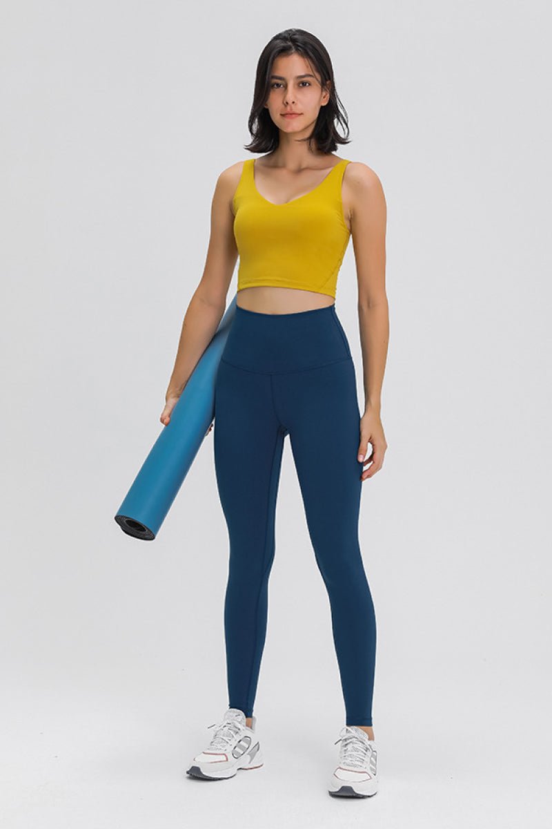Woman wearing a yellow tank top and blue leggings holding a yoga mat on a white background