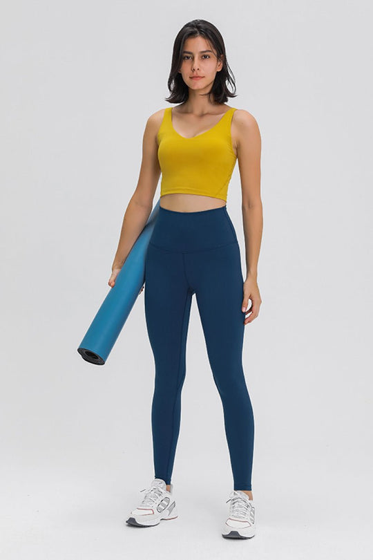 Woman wearing a yellow tank top and blue leggings holding a yoga mat on a white background