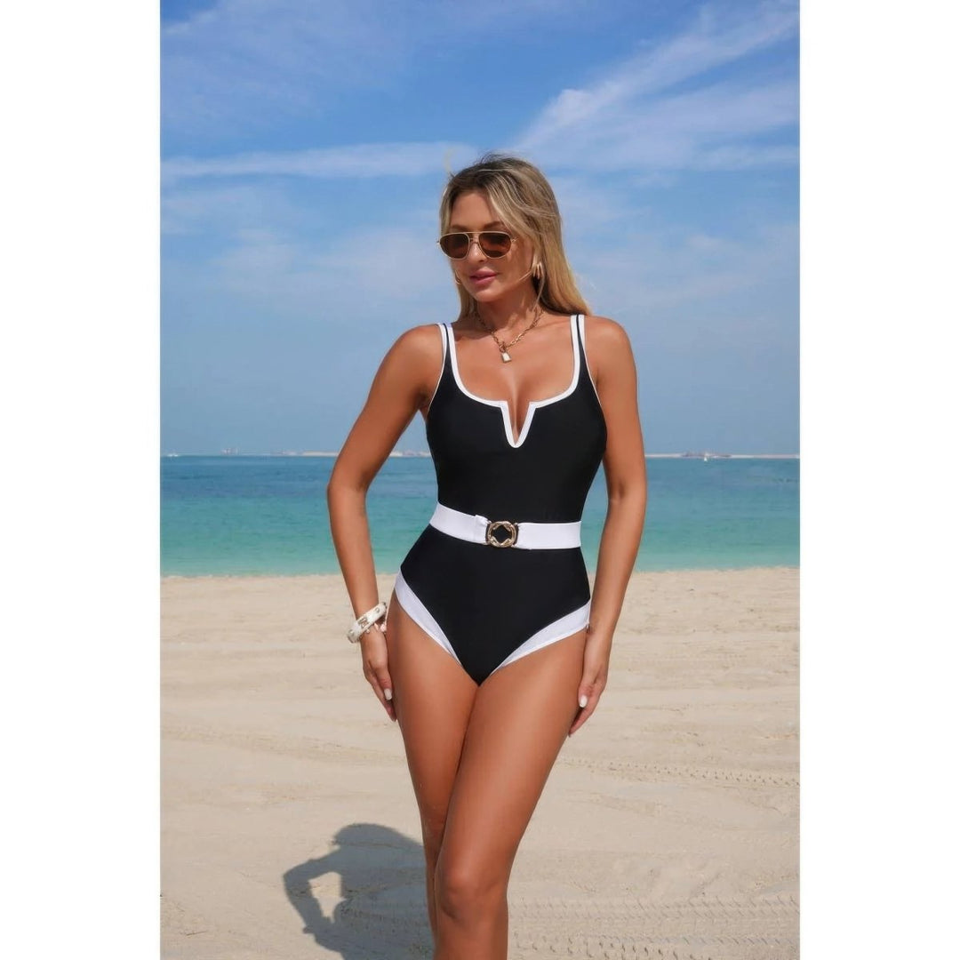 A woman modeling a one-piece swimsuit on a sandy beach with the ocean and blue sky in the background. The swimsuit is black with white contrast trim and a white belt with a gold buckle.