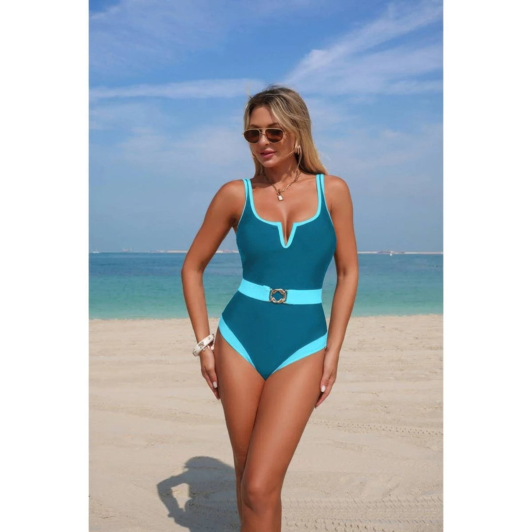 A woman modeling a teal and light blue one-piece swimsuit with a matching belt on a sandy beach with the ocean and blue sky in the background.