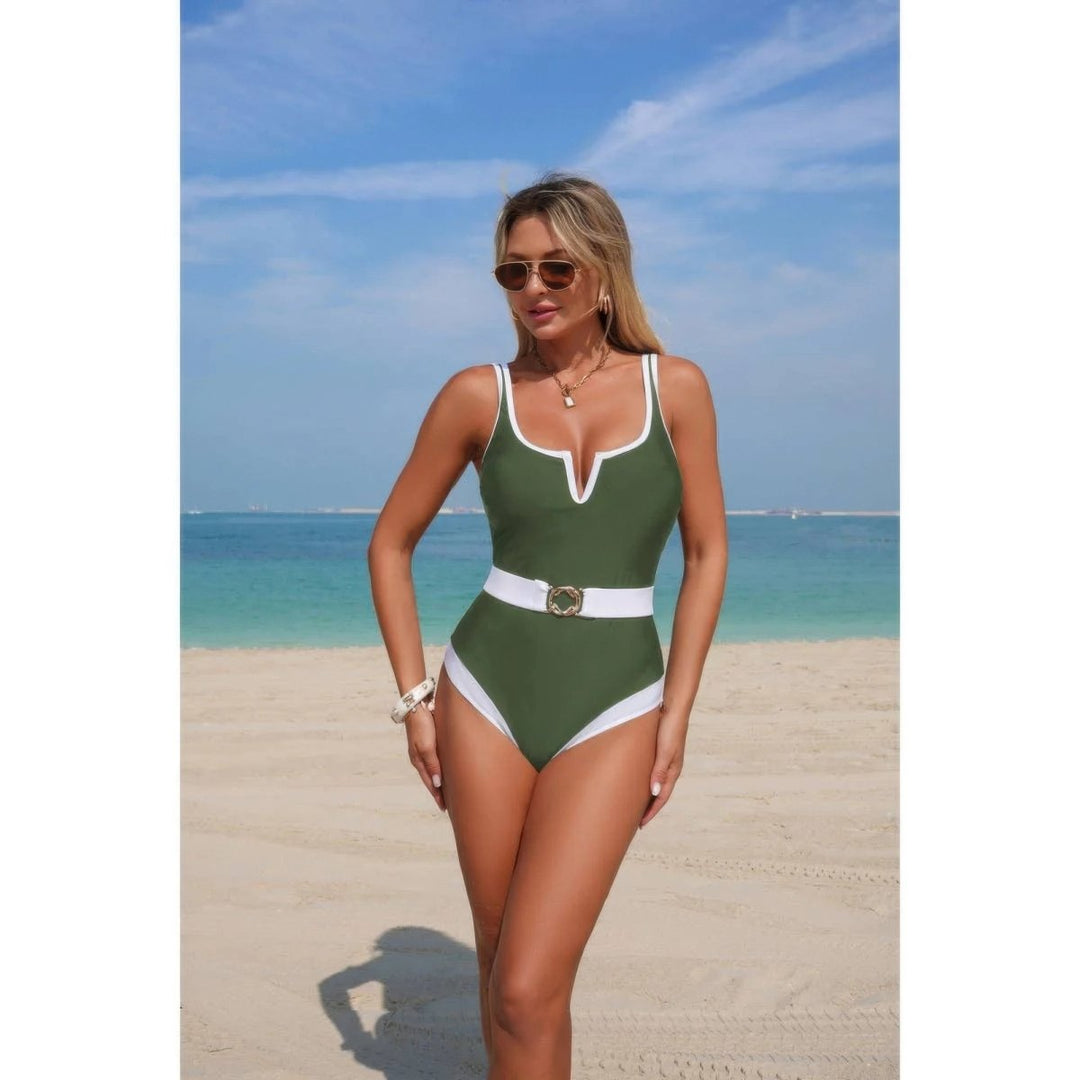A woman modeling a green one-piece swimsuit with white trim and a white belt with a gold buckle, standing on a sandy beach with the ocean and blue sky in the background.