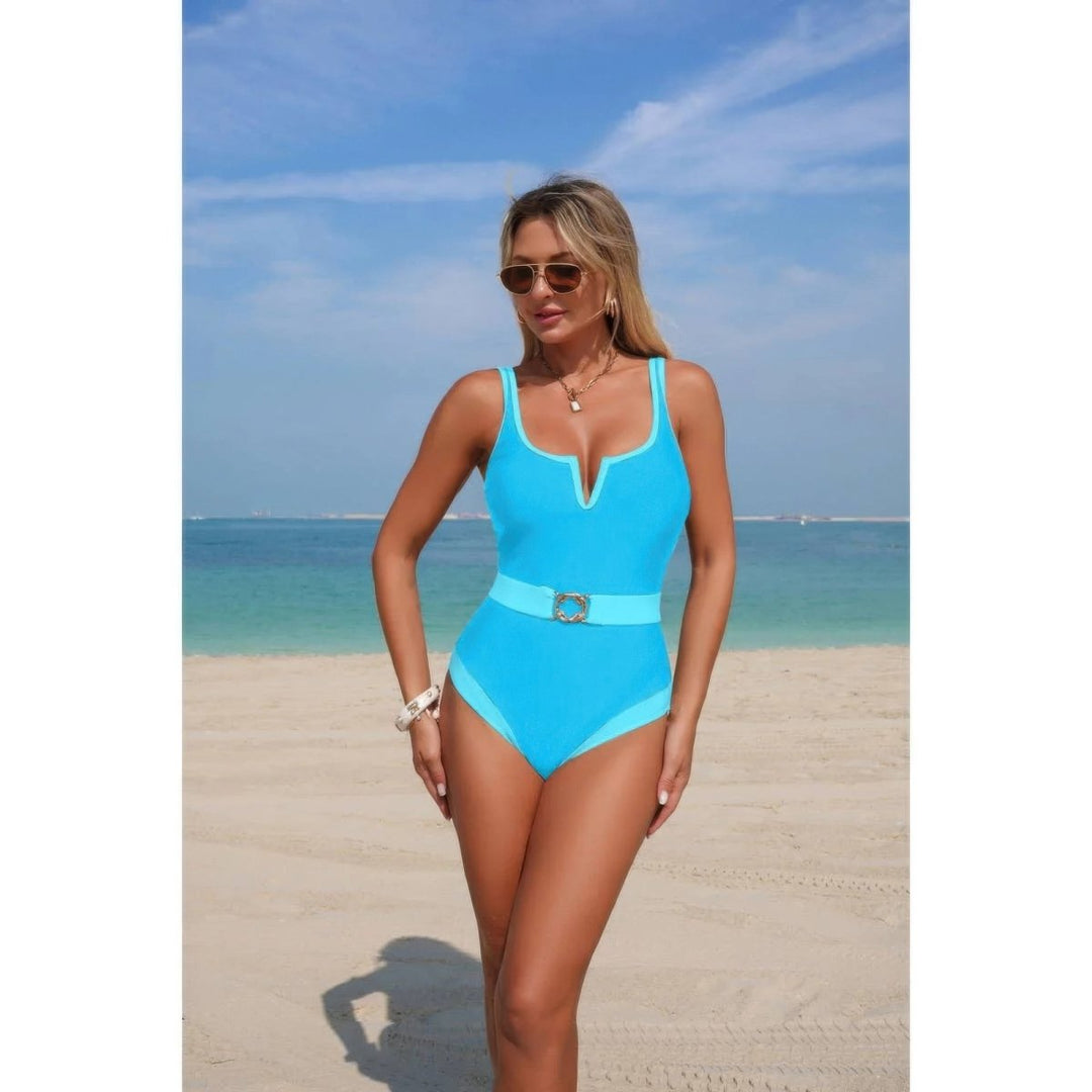A woman modeling a bright turquoise one-piece swimsuit with a matching belt and silver buckle on a sandy beach with the ocean and blue sky in the background.