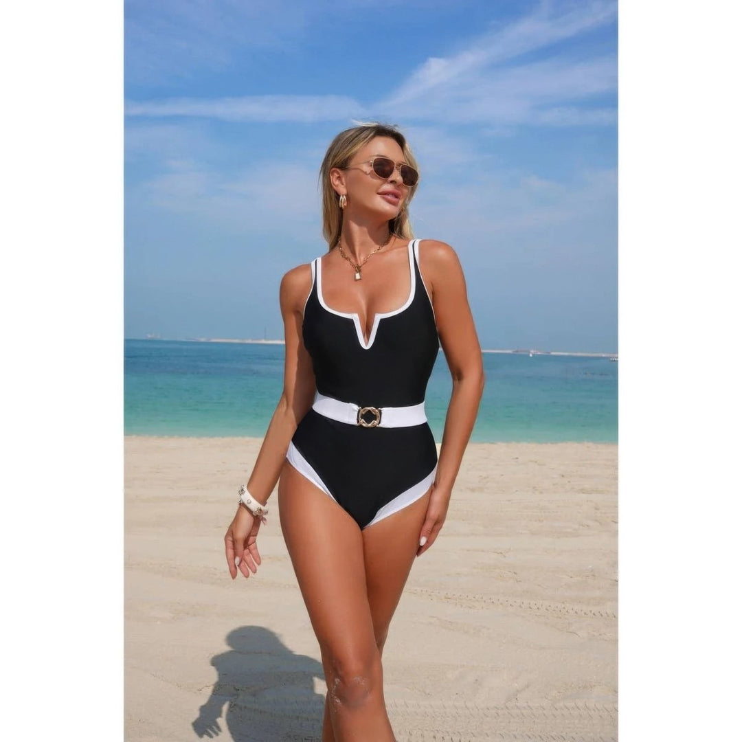 A woman wearing a black one-piece swimsuit with white trim and a white belt stands on a sandy beach with the ocean and blue sky in the background.