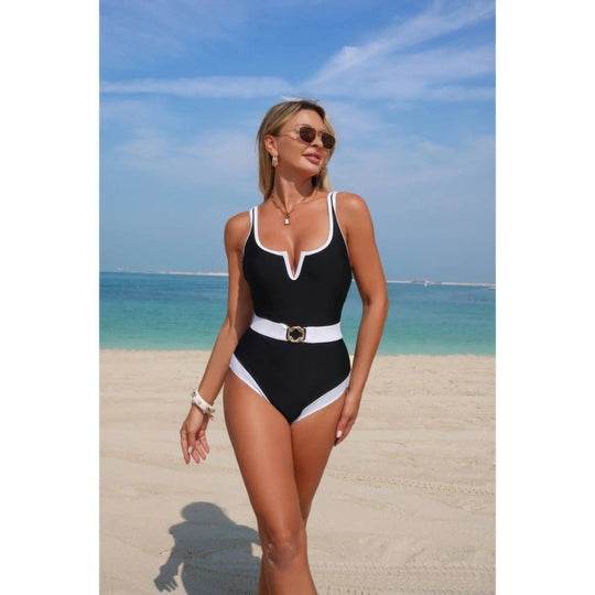 A woman wearing a black one-piece swimsuit with white trim and a white belt stands on a sandy beach with the ocean and blue sky in the background.
