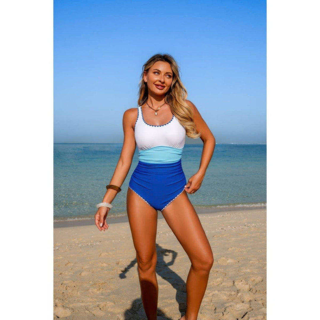 A woman modeling a two-piece swimsuit on a sandy beach with the ocean and blue sky in the background. The swimsuit features a white scoop-neck top with blue trim, a light blue midsection band, and royal blue high-waisted bottoms with white trim.
