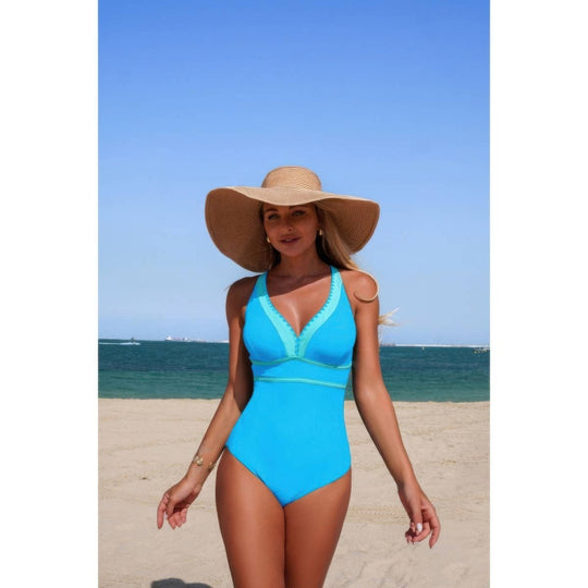 A woman wearing a bright turquoise one-piece swimsuit and a large beige straw sun hat stands on a sandy beach with the ocean and blue sky in the background.