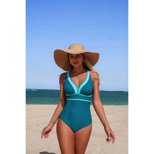 A woman wearing a teal one-piece swimsuit with light blue trim and a large beige straw sun hat, standing on a sandy beach with the ocean and blue sky in the background.