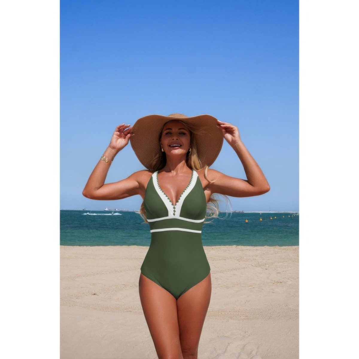 A woman on a sandy beach wearing an olive green one-piece swimsuit with white trim and a large brown sun hat, with the blue ocean and sky in the background.