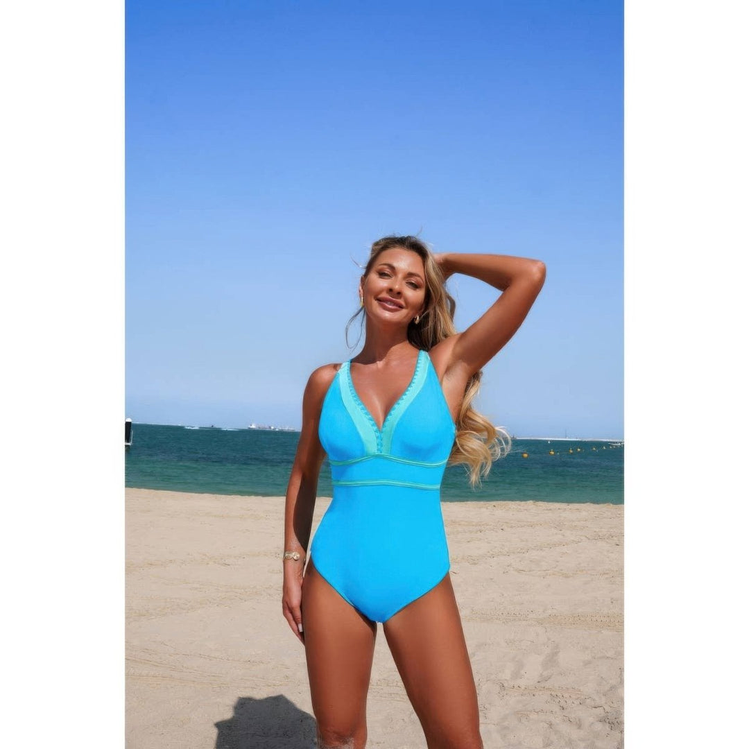 A woman modeling a bright turquoise blue one-piece swimsuit on a sandy beach with the ocean and blue sky in the background.