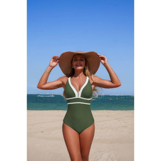 A woman on a sandy beach wearing an olive green one-piece swimsuit with white trim and a large brown sun hat, with the blue ocean and sky in the background.