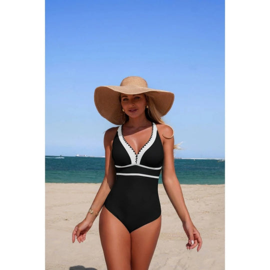 Woman in a black swimsuit with white trim and a wide-brimmed hat on a beach.