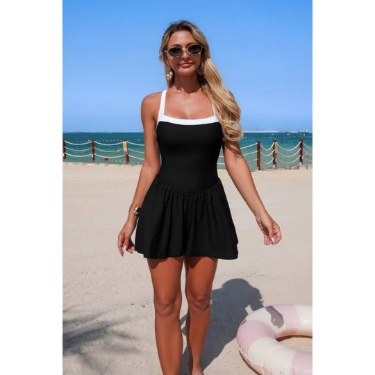 A woman wearing a black one-piece swimsuit with a white trim stands on a sandy beach with the ocean and blue sky in the background.