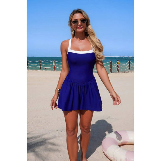 A woman wearing a royal blue one-piece swimsuit with a white trim stands on a sandy beach with the ocean and blue sky in the background.