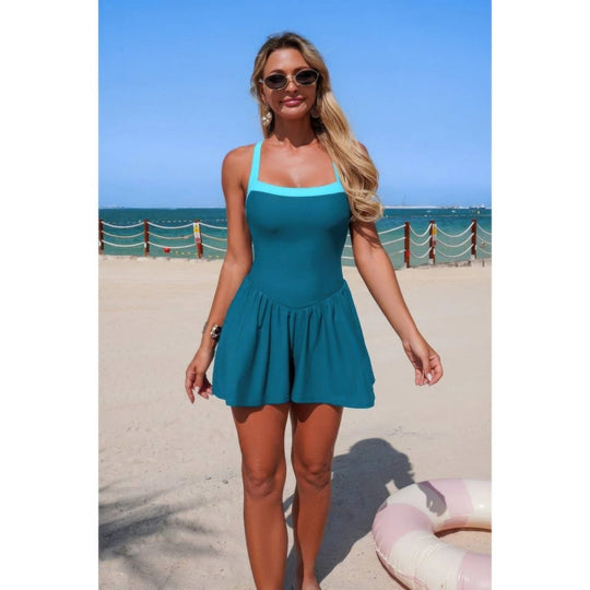 A woman wearing a teal one-piece swimsuit stands on a sandy beach with the ocean and blue sky in the background.