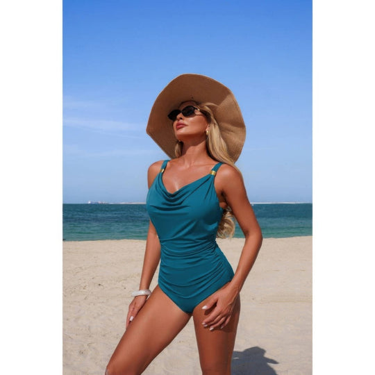 Woman in a teal swimsuit and wide-brimmed hat on a beach with clear blue sky.