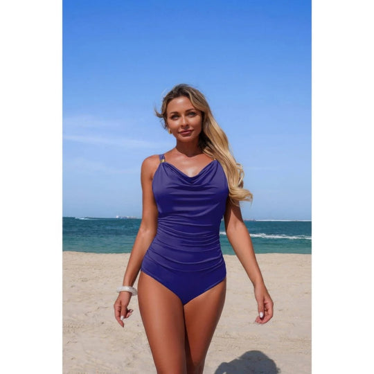 A woman wearing a solid purple one-piece swimsuit stands on a sandy beach with the ocean and blue sky in the background.