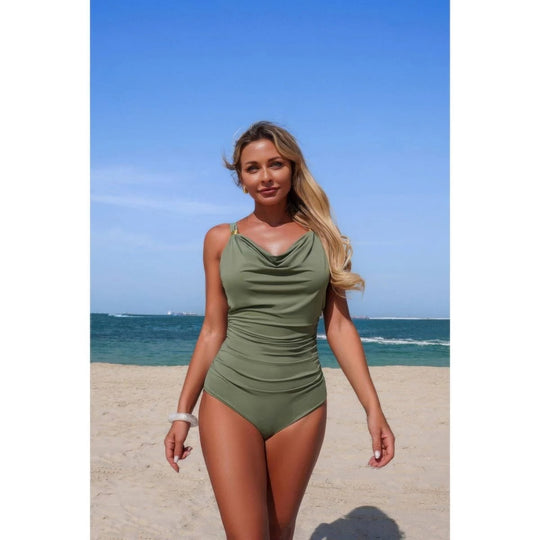 A woman modeling an olive green one-piece swimsuit on a sandy beach with the ocean and blue sky in the background.