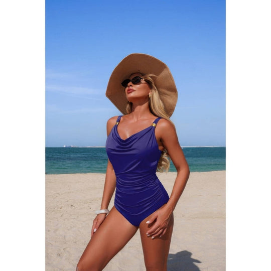 A woman posing on a sandy beach wearing a royal blue one-piece swimsuit, a large brown sun hat, and sunglasses, with the ocean and blue sky in the background.