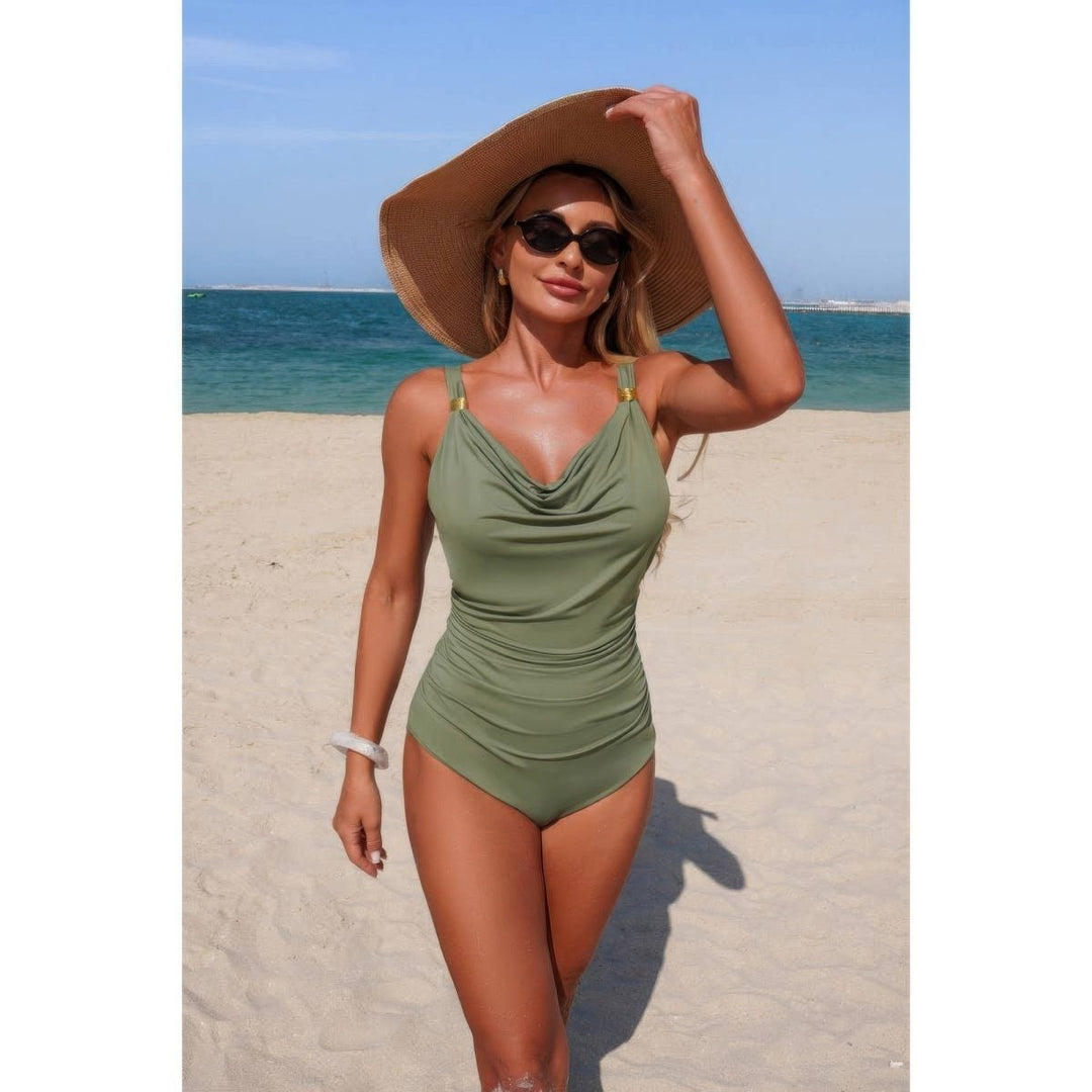 A woman standing on a sandy beach wearing a one-piece olive green swimsuit, a large brown sun hat, and sunglasses, with the ocean and blue sky in the background.