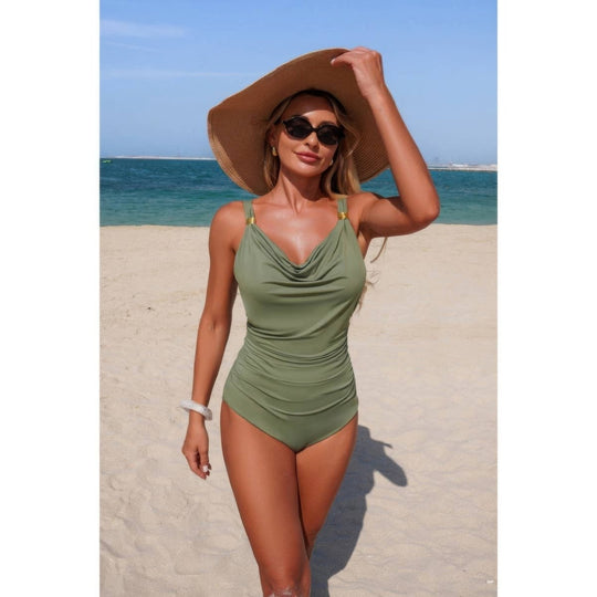A woman standing on a sandy beach wearing a one-piece olive green swimsuit, a large brown sun hat, and sunglasses, with the ocean and blue sky in the background.