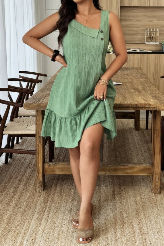 A woman wearing a sage green sleeveless crinkle-textured dress with a ruffled hem, standing in a rustic dining room next to a wooden table.