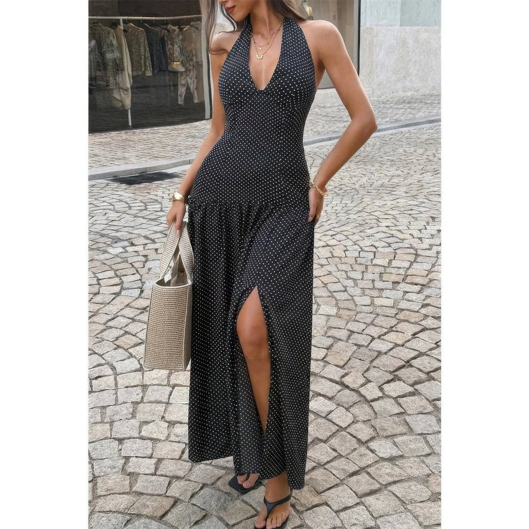 A woman wearing a sleeveless black maxi dress with a small white polka dot pattern, a deep V-neckline, and a high side slit, standing on a cobblestone street and holding a woven tote bag.