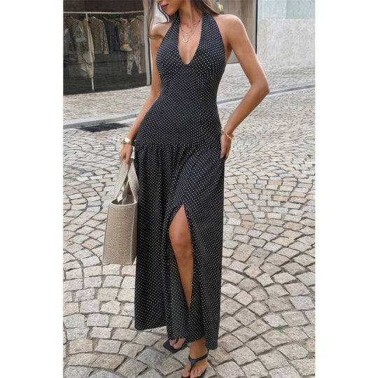 A woman wearing a sleeveless black maxi dress with a small white polka dot pattern, a deep V-neckline, and a high side slit, standing on a cobblestone street and holding a woven tote bag.