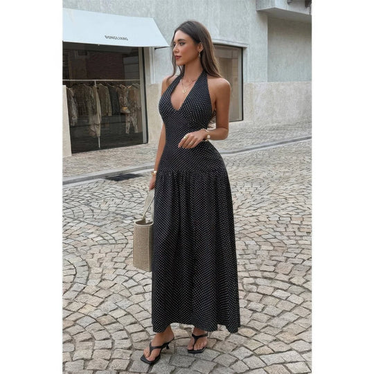 A woman stands on a cobblestone street wearing a sleeveless, halter-neck maxi dress with a small white polka dot pattern on a black base. She is holding a beige woven handbag and wearing black strappy heels.