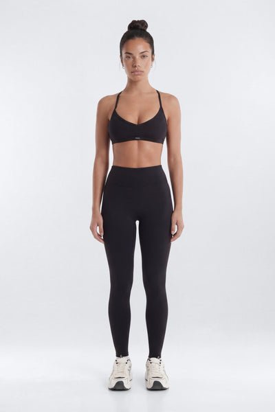 A woman stands facing forward in a black sports bra and black high-waisted leggings, paired with white sneakers, against a plain light gray studio background.