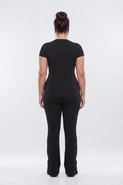A woman viewed from the back wearing a black short-sleeve top and matching black pants against a plain light gray studio background.