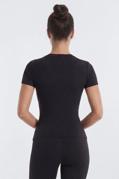 Rear view of a woman with her hair in a bun wearing a black short-sleeve crewneck top and black leggings against a light gray studio background.
