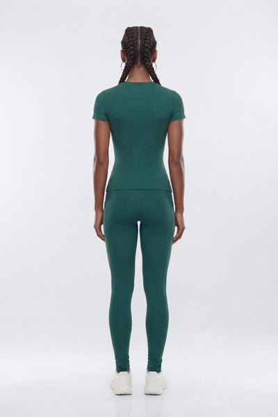 A woman viewed from the back wearing a matching dark green short-sleeve top and leggings set, paired with white sneakers, against a light gray studio background.