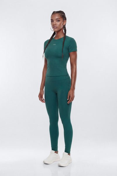 A woman modeling a matching dark teal green athletic set consisting of a short-sleeve crewneck top and high-waisted leggings, paired with white sneakers, against a light gray studio background.