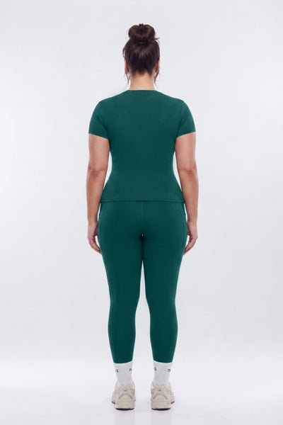 A woman viewed from the back wearing a matching dark green short-sleeve top and leggings set, with white socks and beige sneakers, against a plain light gray studio background.