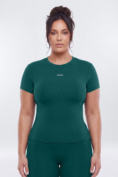 A woman modeling a dark green short-sleeve athletic top with a small white logo on the chest, paired with matching green leggings, against a light gray studio background.
