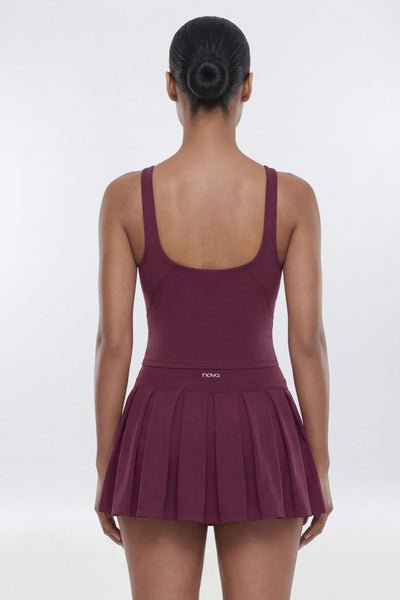 A woman shown from the back wearing a sleeveless athletic dress with a scoop neckline and a pleated skirt, in a deep plum-burgundy color. The brand name &