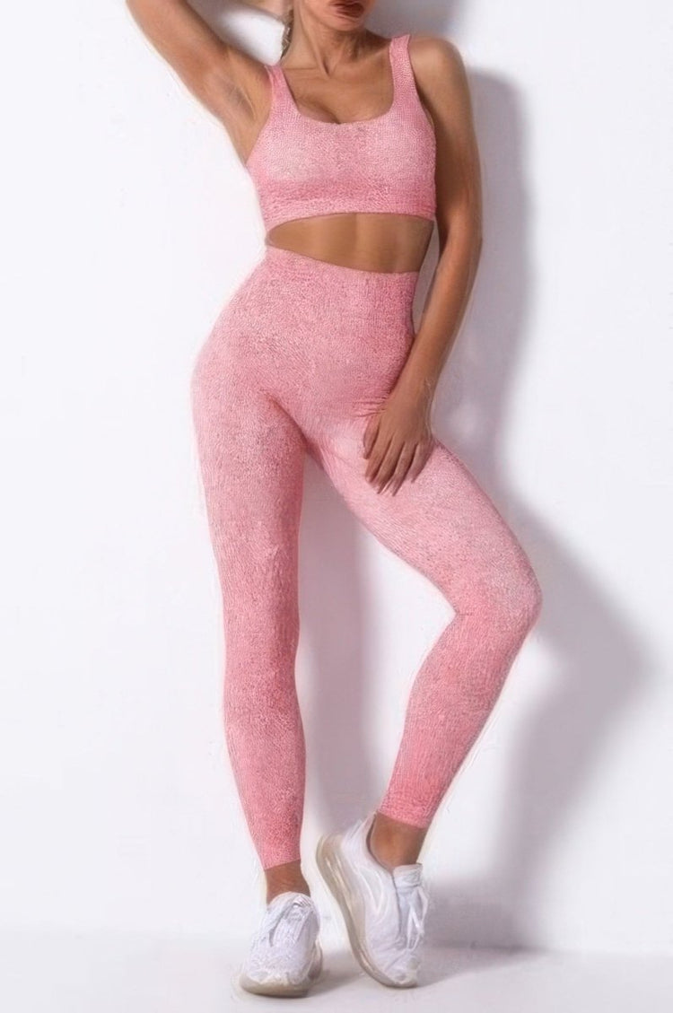 A woman modeling a matching pink activewear set consisting of a sports bra and high-waisted leggings with a subtle textured pattern, paired with white sneakers, against a white wall.