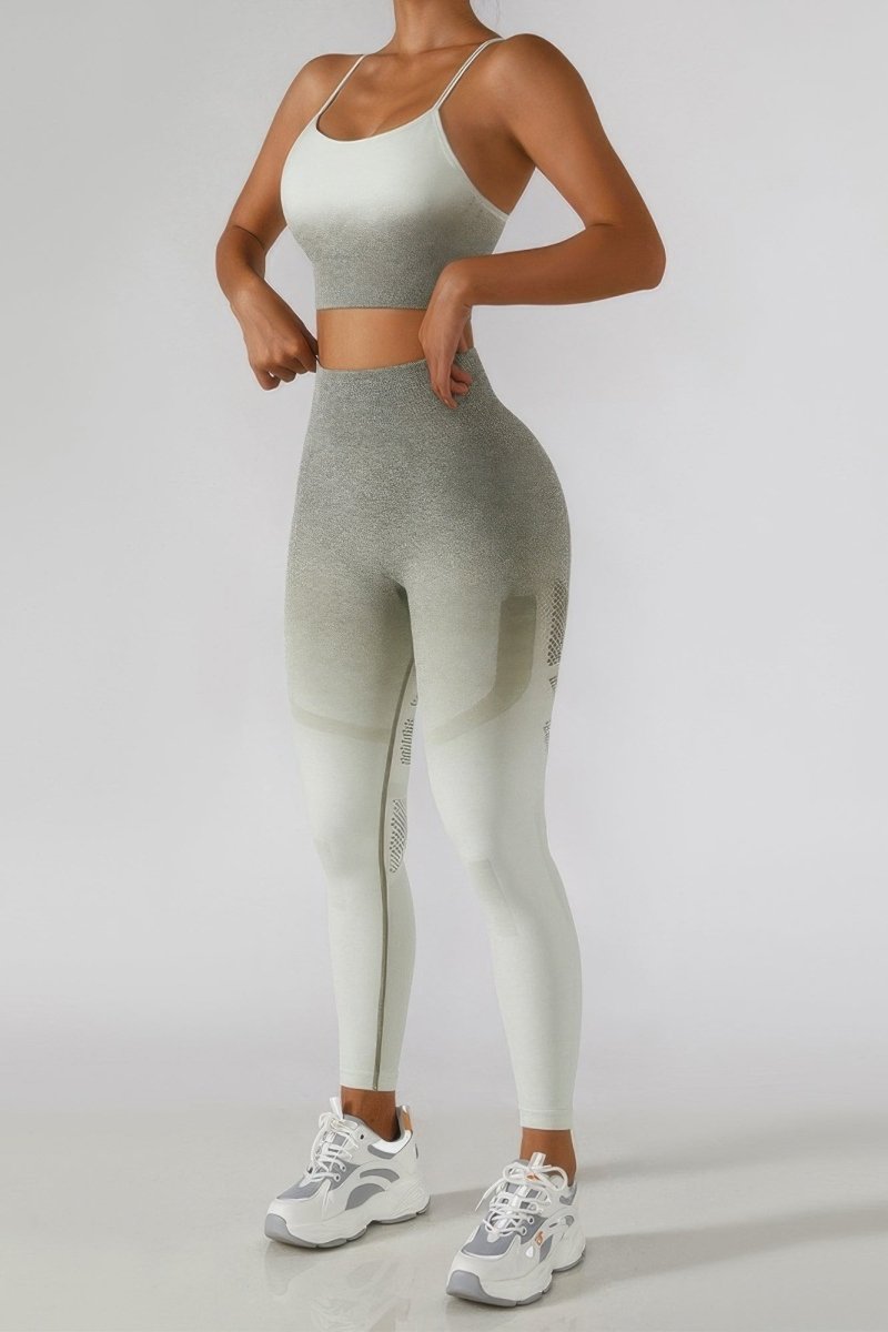 A woman modeling a matching two-piece activewear set consisting of a light gray sports bra and high-waisted leggings with a gradient fade to off-white, paired with white and gray sneakers, against a plain light gray studio background.